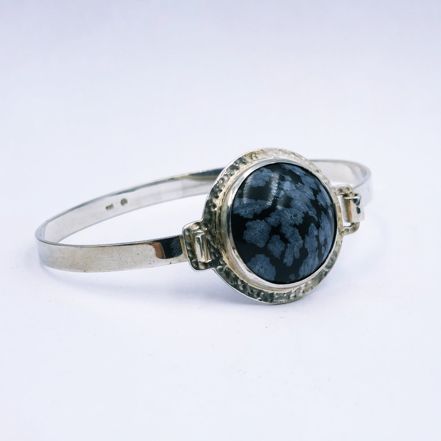 Sterling silver oval bangle with clasp, featuring a 25mm snowflake obsidian set in a hammered round tube bezel, handmade with 74mm diameter.