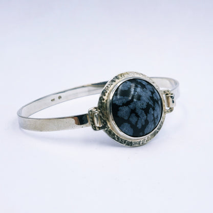 Sterling silver oval bangle with clasp, featuring a 25mm snowflake obsidian set in a hammered round tube bezel, handmade with 74mm diameter.